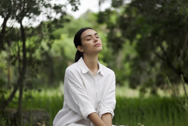 A photo of a woman sitting outdoors in a serene setting, with greenery all around. She is wearing a white shirt and has her eyes closed. Her posture is straight, and she is sitting on a rock. The background is filled with trees and greenery. The lighting is soft.