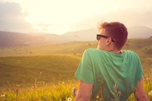 A person wearing sunglasses and a green shirt sits in a grassy field, facing the sunset over rolling hills.