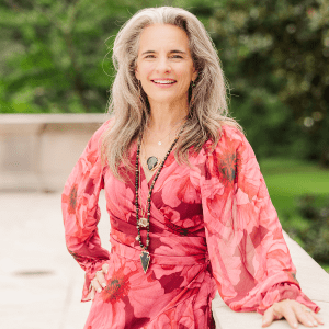 A woman with long gray hair wearing a red floral dress and layered necklaces stands outdoors, smiling at the camera.