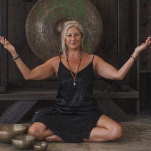 A woman in a black dress sits cross-legged on the floor with arms raised, surrounded by singing bowls, in front of a large gong.