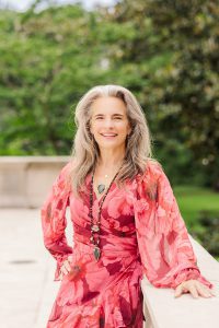 A woman with long gray hair wearing a red floral dress stands outdoors, leaning on a stone railing, with greenery in the background.