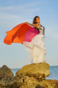 A woman stands on rocks by the sea, holding a flowing orange and pink fabric behind her, with a blue sky in the background.