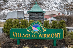 A green sign with "Village of Airmont" and "Welcome to the Village, Founded 1991" stands in front of bushes and a small gazebo-like structure. Cars and a Walgreens are visible in the background.