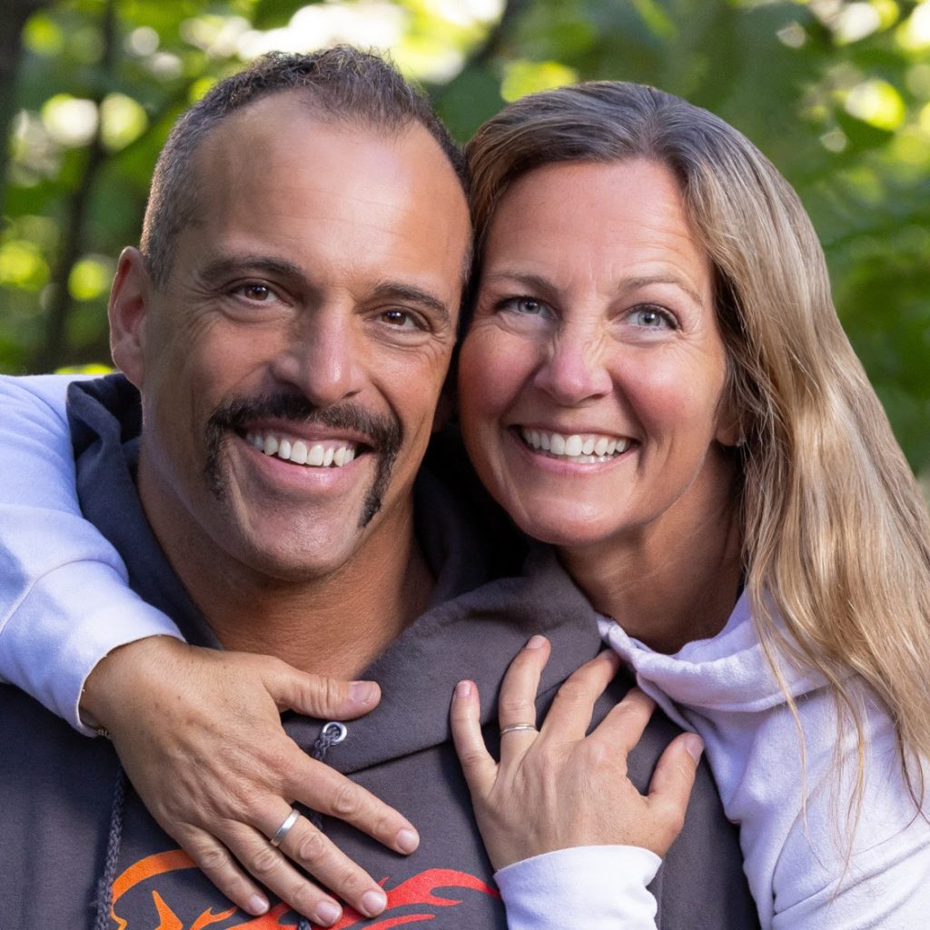 A man and woman smiling outdoors, with the woman embracing the man from behind. Both are wearing casual hoodies and rings, with green foliage in the background.
