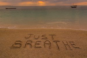 The words "JUST BREATHE" are written in sand on a beach with calm water, two boats, and a cloudy sky in the background.