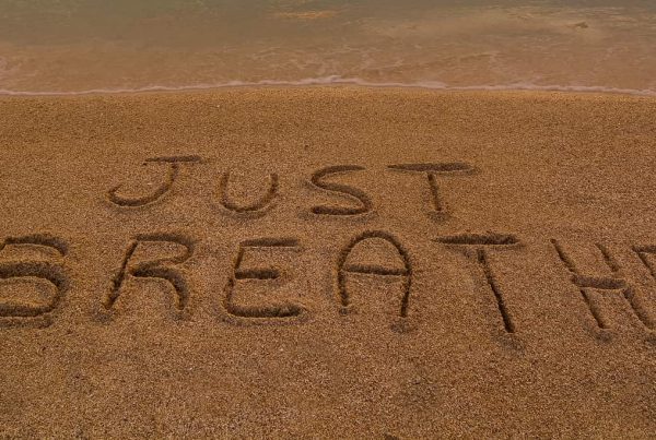 The words "JUST BREATHE" are written in the sand near the water's edge on a beach.