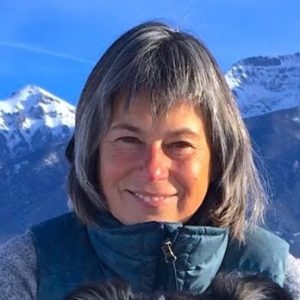 A woman with gray hair and a blue vest smiles outdoors with snow-covered mountains in the background.