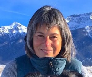 A woman with short gray hair and a blue jacket stands outdoors with snow-covered mountains in the background.