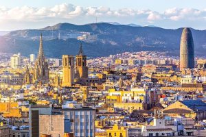 A panoramic view of Barcelona featuring historic buildings, the Sagrada Família, Torre Glòries, and distant mountains under a partly cloudy sky.