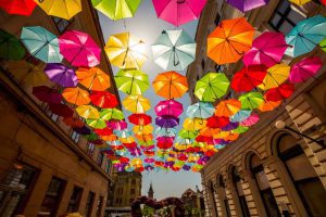 Colorful umbrellas hang suspended above a pedestrian street between two buildings, creating a vibrant canopy under a sunny sky.