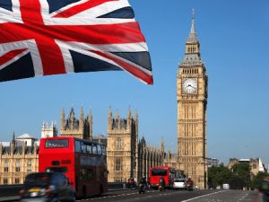A Union Jack flag flies near Big Ben and the Houses of Parliament in London, with red double-decker buses on the street below under a clear blue sky.