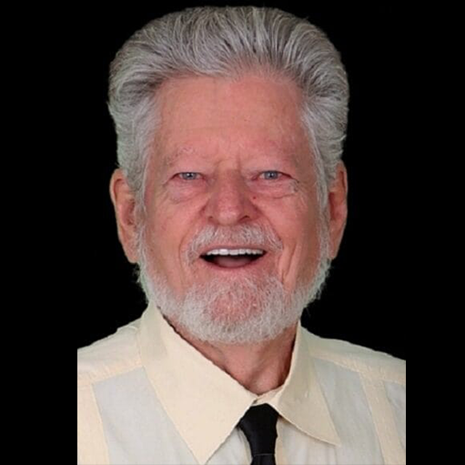 An older man with gray hair and a beard is smiling, wearing a light-colored shirt and a black tie, against a black background.