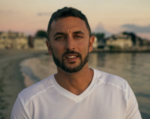 A man with short dark hair and a trimmed beard stands on a beach at sunset, wearing a white shirt with buildings and water in the background.