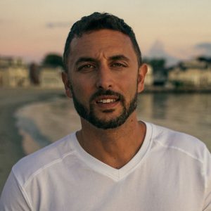 A man with short dark hair and a beard wearing a white shirt stands on a beach at sunset, with buildings and water in the background.