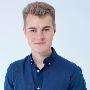 A young man with short blond hair wearing a dark blue button-up shirt stands against a plain light background, looking at the camera.