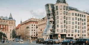 Street view of the Dancing House in Prague, featuring modern curved architecture alongside traditional buildings, with cars and pedestrians in the foreground.