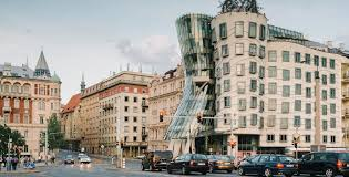 Street view of the Dancing House in Prague, featuring modern curved architecture alongside traditional buildings, with cars and pedestrians in the foreground.