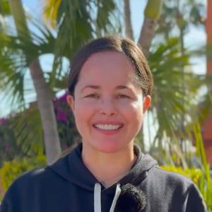 A woman wearing a black hoodie smiles outdoors with palm trees and greenery in the background.