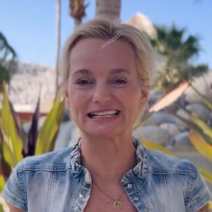 A woman with short blonde hair wearing a denim jacket stands outdoors, smiling at the camera with palm trees and rocks in the background.