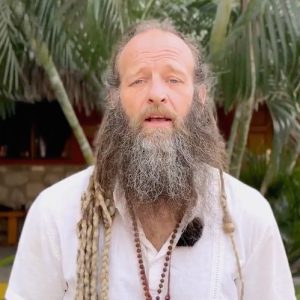 A man with long dreadlocks and a full beard stands outdoors in front of palm trees, wearing a white shirt and a beaded necklace.