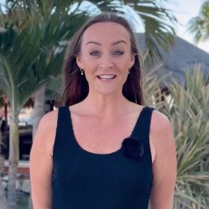 A woman with long brown hair in a sleeveless black top stands outdoors in front of palm trees, smiling at the camera.