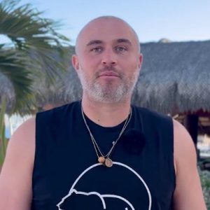 A man with a shaved head and beard stands outdoors in front of a thatched-roof structure, wearing a sleeveless black shirt and two necklaces.