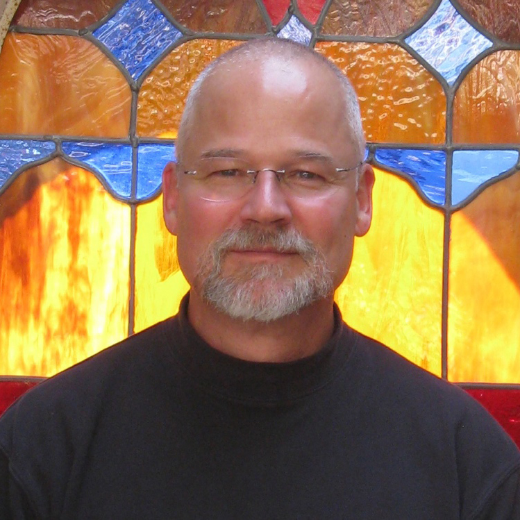 A middle-aged man with glasses and a gray beard is standing in front of a stained glass window, wearing a black shirt.