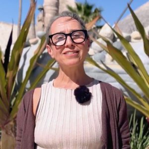 A woman with glasses stands outdoors in front of tall plants, smiling at the camera. She wears a sleeveless top and a brown cardigan.