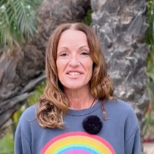 A woman with long brown hair and a nose ring stands outdoors in front of a tree, wearing a gray sweatshirt with a rainbow graphic and a clip-on microphone.