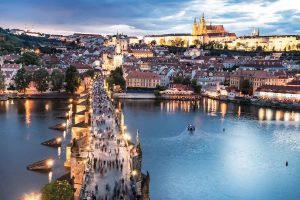 Aerial view of the Charles Bridge spanning the Vltava River, with Prague Castle illuminated in the background at dusk.