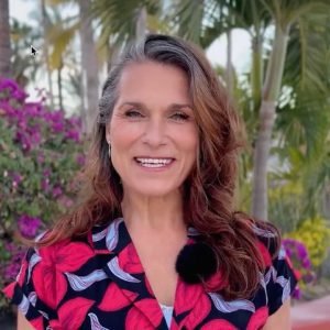 A woman with long brown hair wearing a floral-patterned top stands outdoors, smiling at the camera, with palm trees and purple flowers in the background.