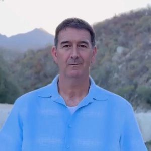 A man in a light blue shirt stands outdoors with mountains and vegetation in the background.