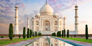 The Taj Mahal, a white marble mausoleum with minarets, stands symmetrically behind a long reflecting pool and manicured gardens at sunset.