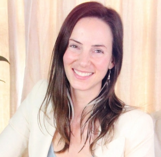 A woman with long brown hair, wearing a light-colored blazer and earrings, smiles while sitting in front of beige curtains.