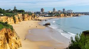 Sandy beach with cliffs, a few people walking along the shore, gentle waves, and a city skyline in the background under a clear sky.