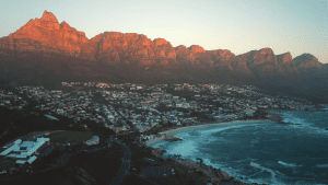 Aerial view of a coastal town with mountains in the background, bathed in warm sunlight, with houses near the shoreline and waves crashing onto the beach.