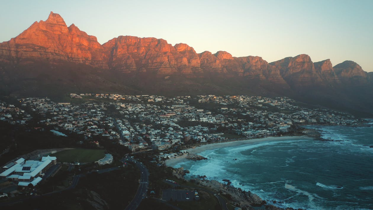 Aerial view of a coastal town with mountains in the background, bathed in warm sunlight, with houses near the shoreline and waves crashing onto the beach.