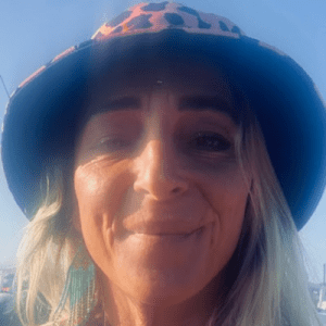 A person with long blonde hair wearing a leopard-print bucket hat and teal earrings smiles at the camera outdoors in bright sunlight.