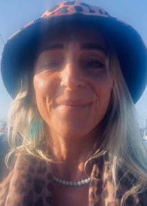 A woman with long blonde hair wears a leopard print hat and scarf, turquoise earrings, and a pearl necklace, smiling outdoors in bright sunlight.