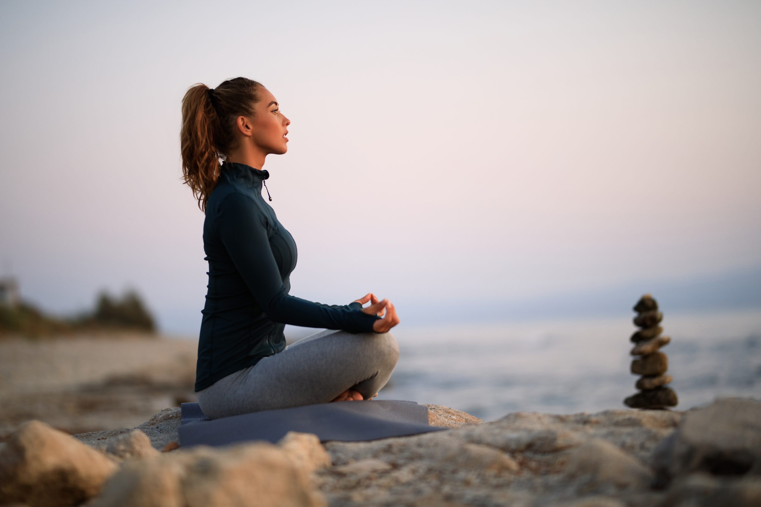 A woman sits in a cross-legged meditation pose on a rock by the ocean, near a small stack of balanced stones.